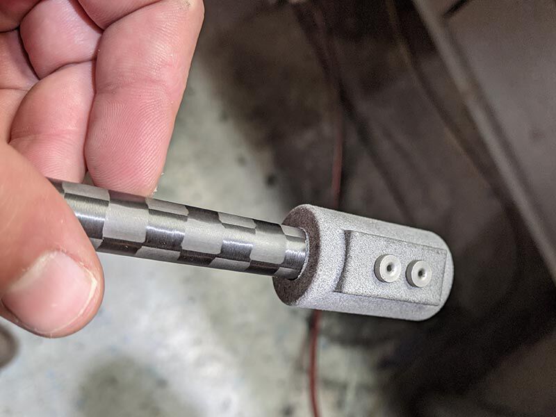 Image shows a fingers holding a small carbon fiber tube with an assembly fixture bonded to the end. The carbon tube has a checkboard surface and metallic grey fixture has two circular inserts.